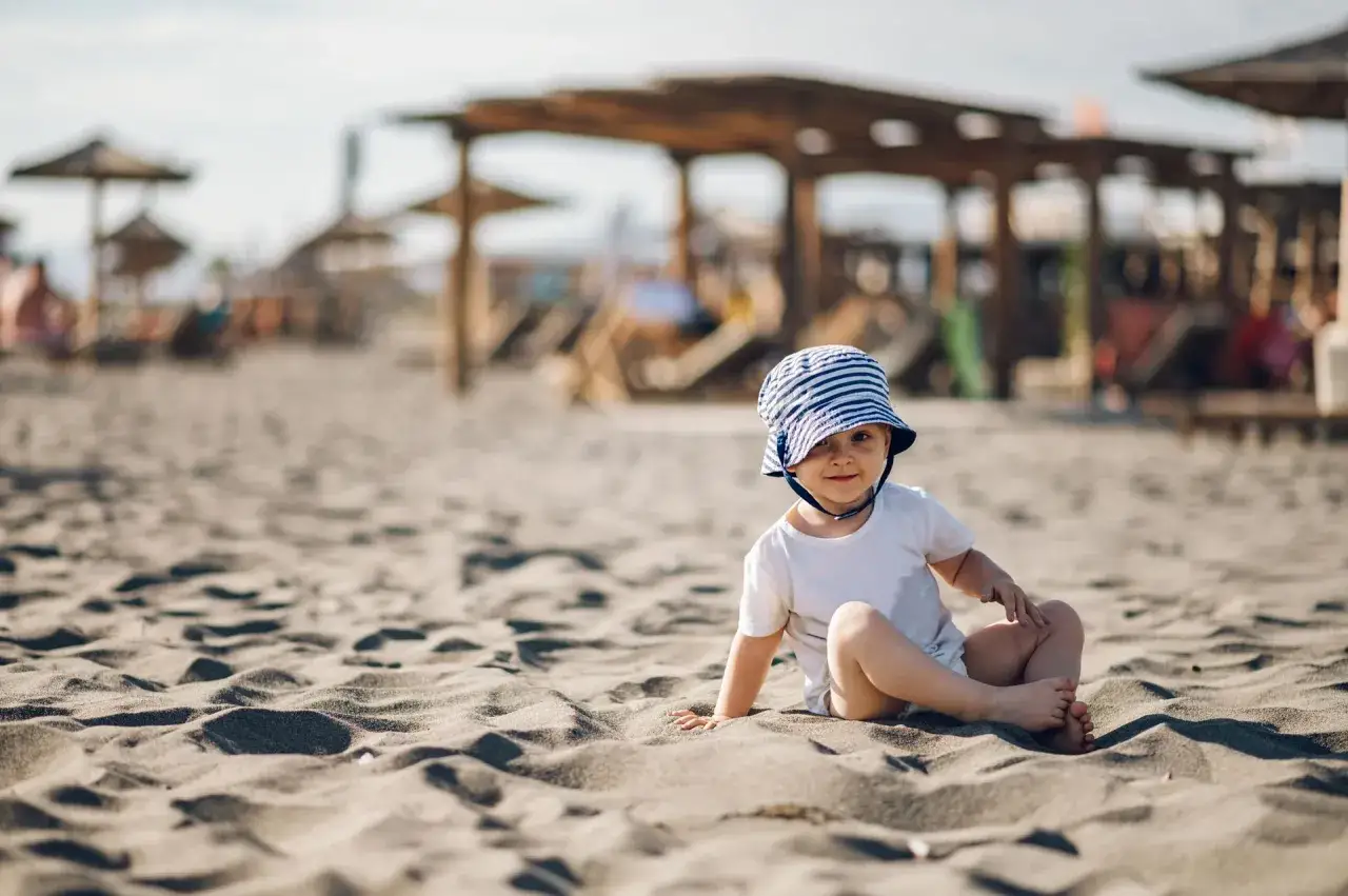 Uroczy maluch w pasiastym kapelusiku siedzi na piaszczystej plaży. Idealne miejsce, gdzie na wakacje z rocznym dzieckiem.