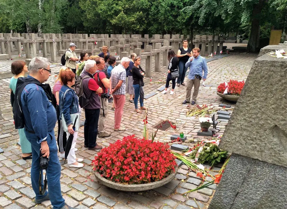Grupa zwiedzających słucha przewodnika przy pomnikach w Warszawie. W tle rzędy krzyży, przed nimi kwiaty i znicze.