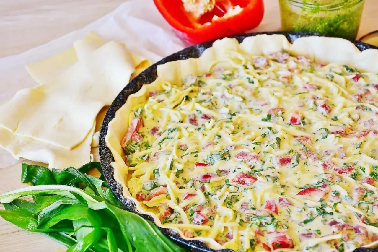 Quiche Lorraine w trakcie przygotowania, z widocznym ciastem, nadzieniem z boczku, sera i zi&oacute;ł, obok papryka i dziki czosnek.
