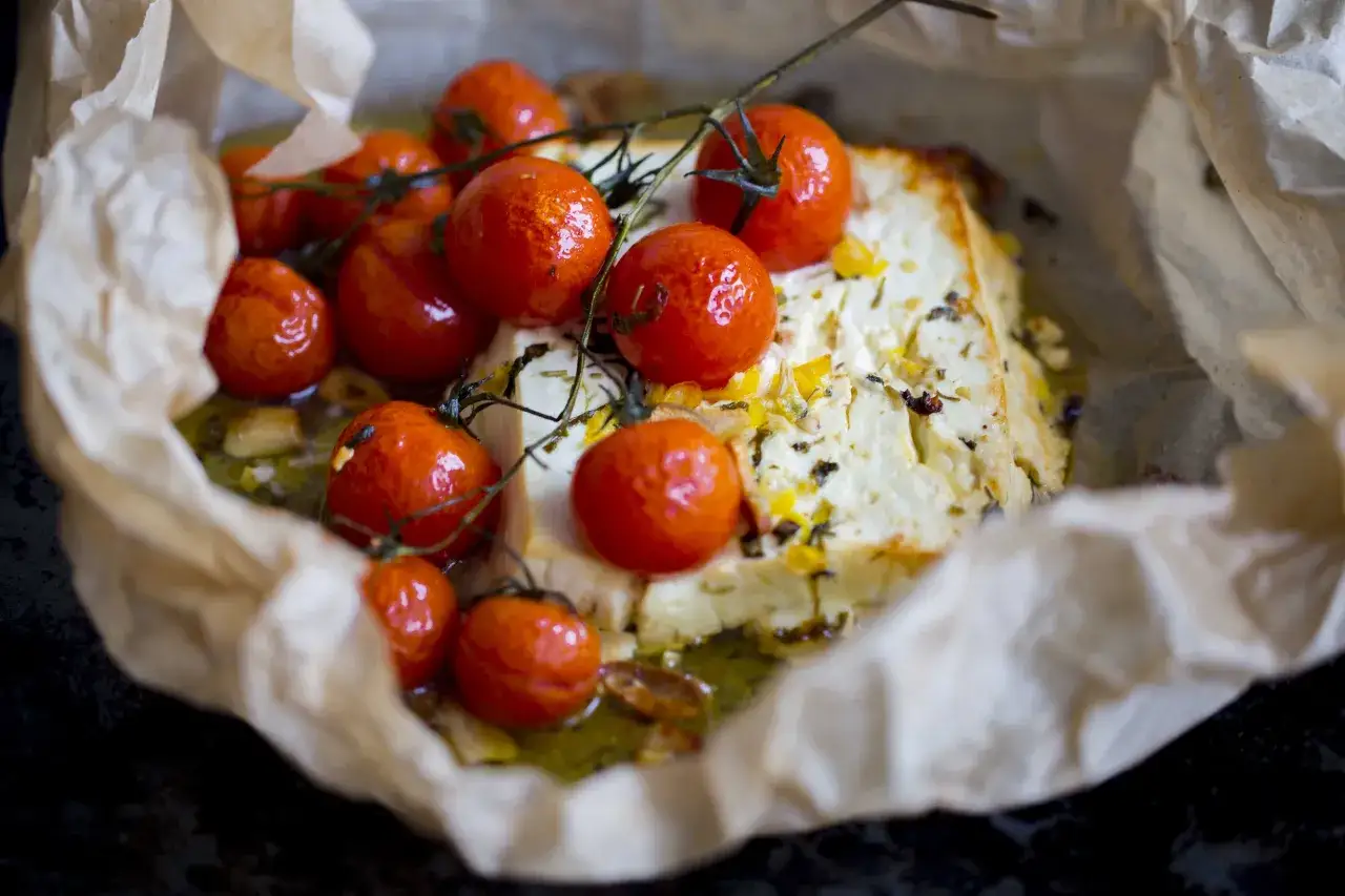 Pyszna feta zapiekana z pomidorkami koktajlowymi, ziołami i oliwą, podana na papierze do pieczenia.