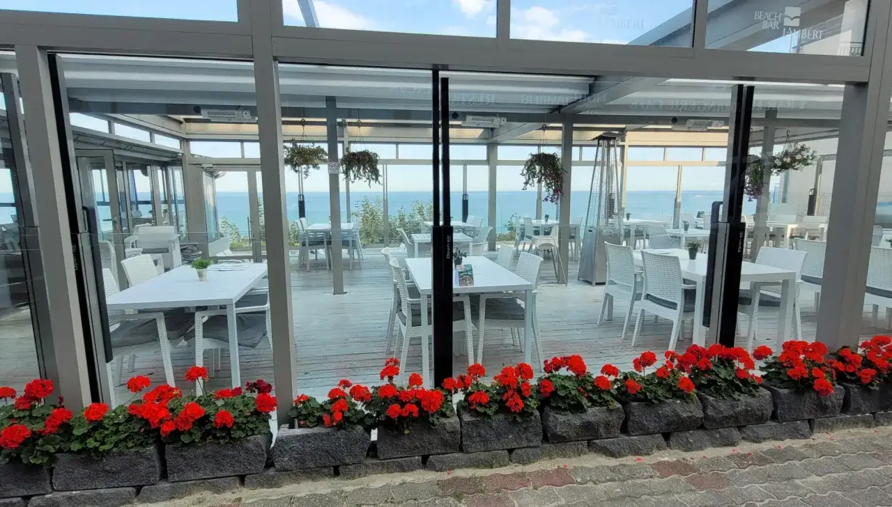 Letnia atmosfera przy plaży. Stoliki i krzesła w przeszklonym ogródku restauracyjnym hotelu Lambert, z widokiem na morze.