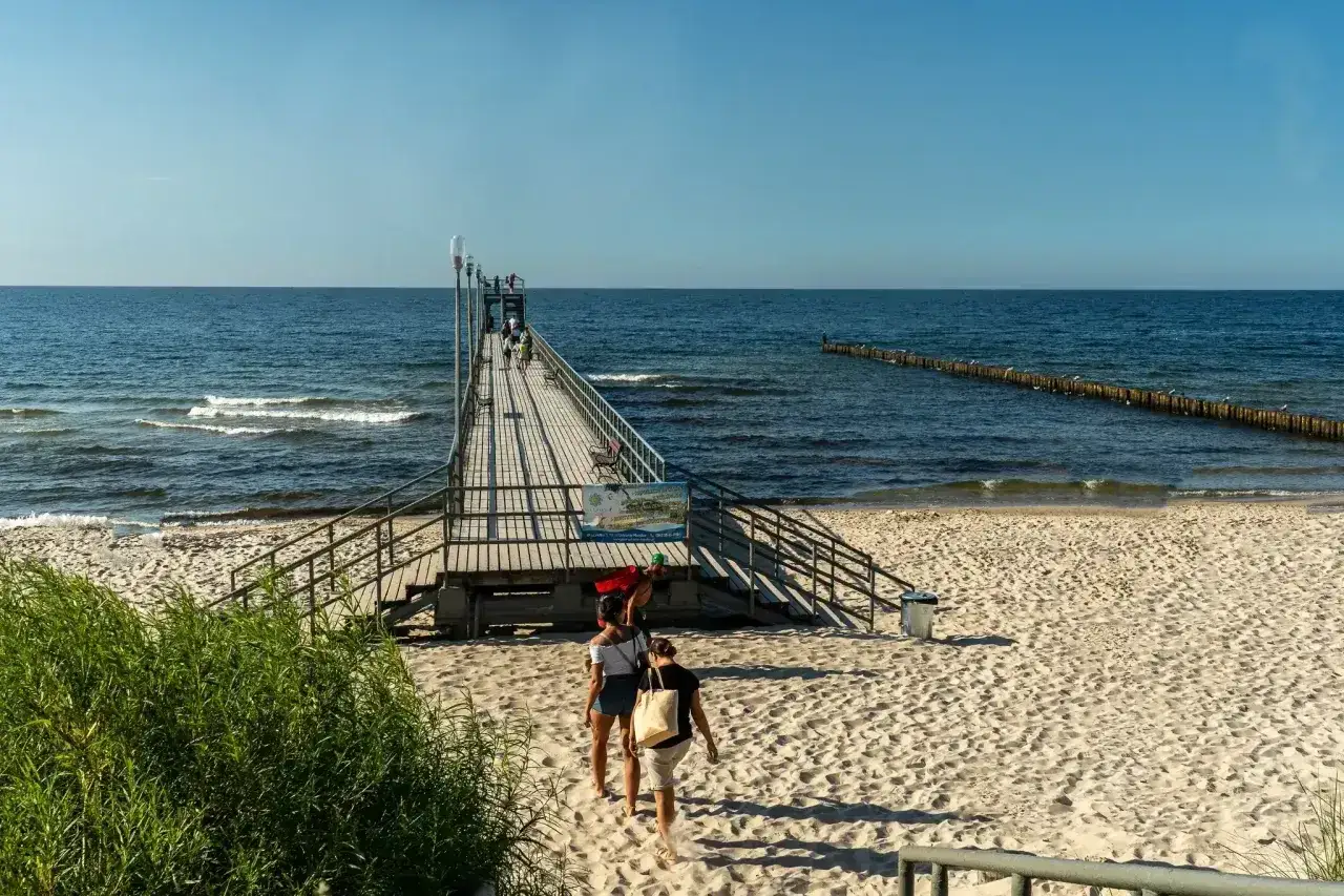 Sianożęty atrakcje: drewniane molo na plaży, ludzie spacerują, morze i błękitne niebo.