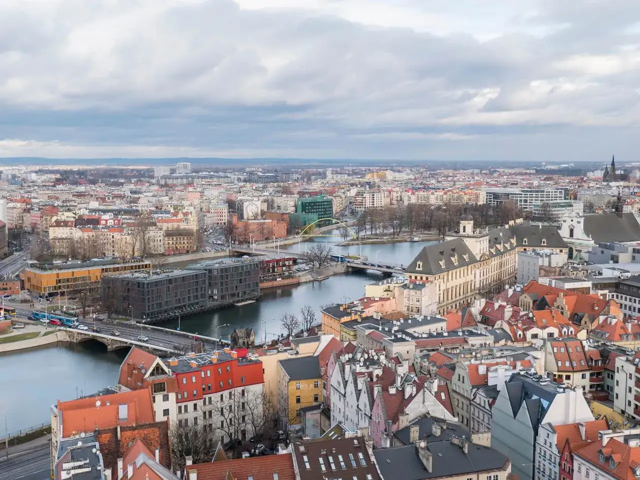 Widok na Wrocław z góry, z rzeką i licznymi budynkami. W oddali widać panoramę miasta, która przypomina widoki z wież widokowych Dolnego Śląska.