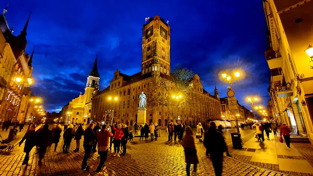 Wieczorne atrakcje Torunia: Rynek Staromiejski z Ratuszem i pomnikiem Kopernika, oświetlony brukowany deptak pełen spacerowiczów.