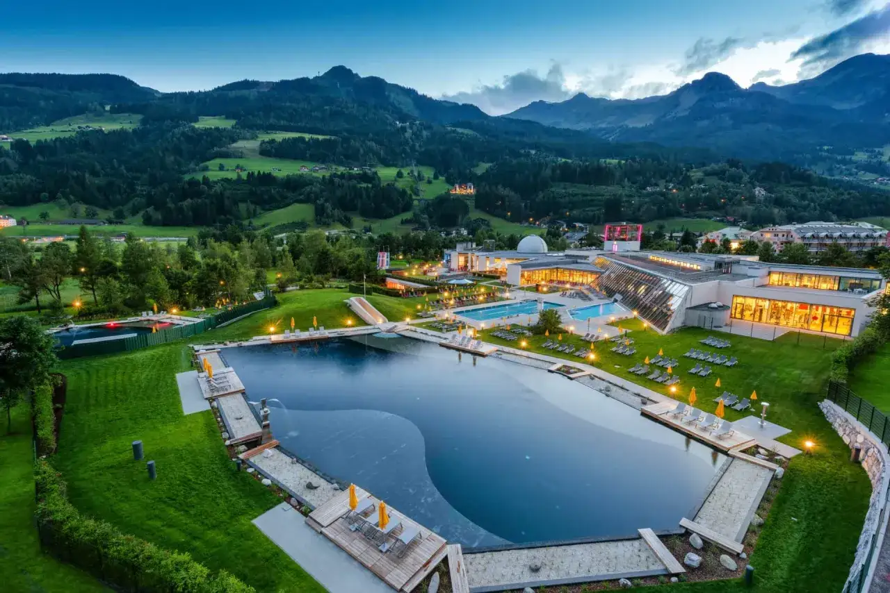 Baseny i nowoczesny kompleks termalny w Bad Gastein, otoczone zielonymi wzg&oacute;rzami i g&oacute;rami, oświetlone wieczornym blaskiem.