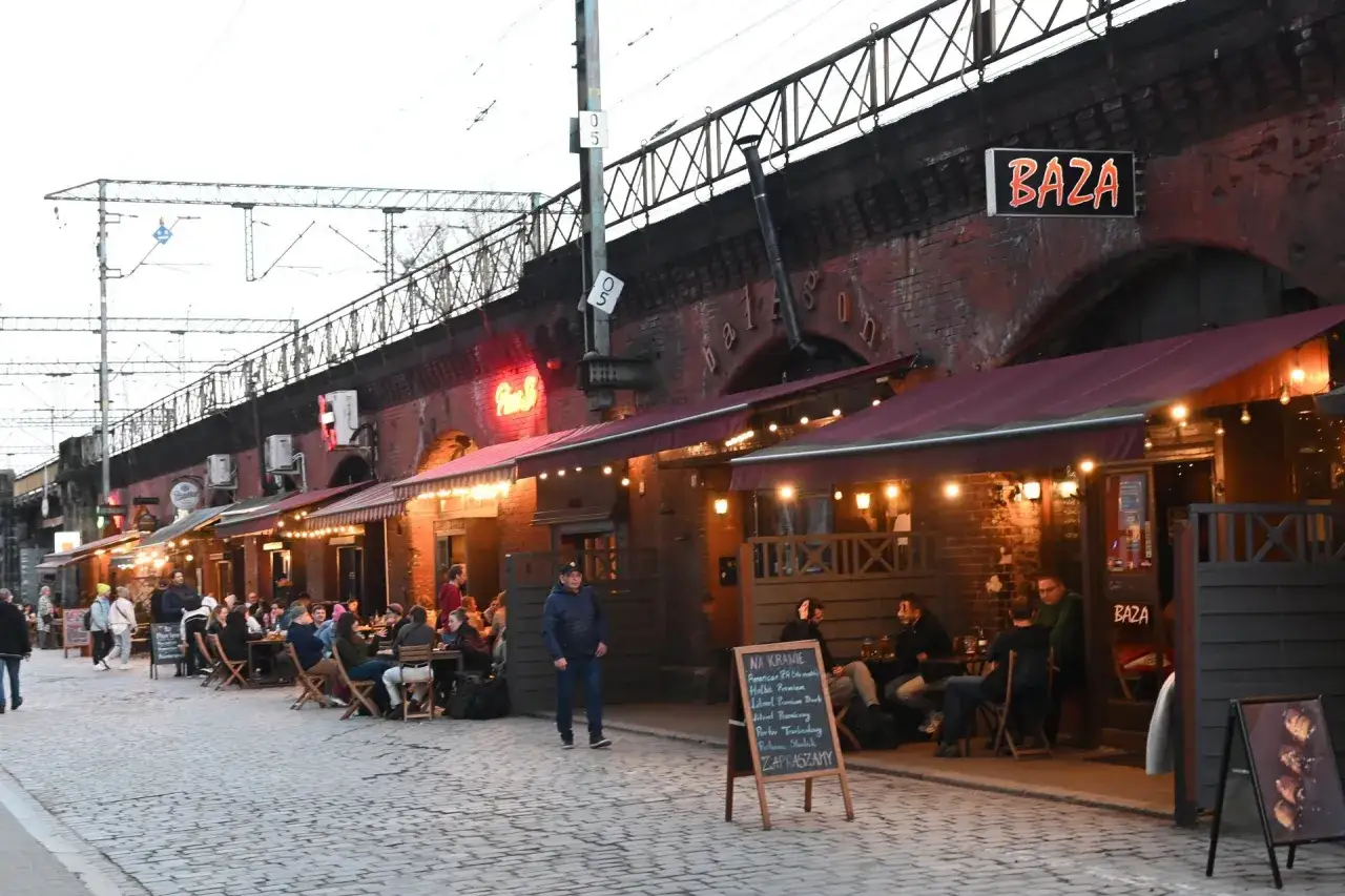 Tętniący życiem nasyp we Wrocławiu, gdzie pod ceglanymi łukami kolejowymi rozsiadły się ogródki kawiarniane. Ludzie siedzą przy stolikach, ciesząc się wieczorem.