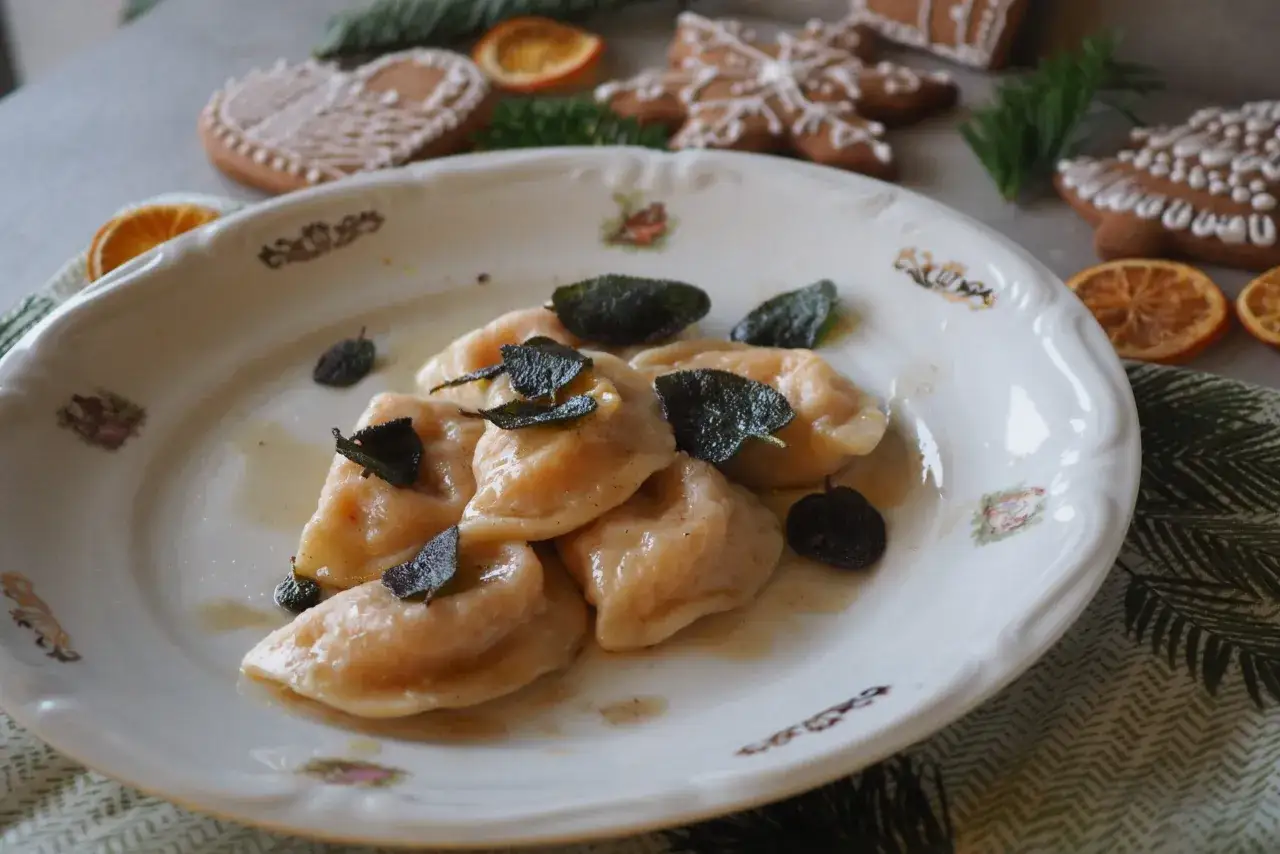 Pyszne pierogi z dynią, polane masłem z szałwią, na talerzu ozdobionym świątecznymi pierniczkami i suszonymi pomarańczami.