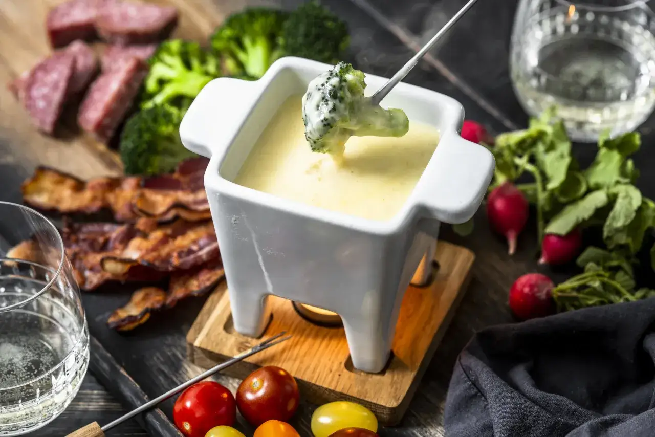 Pyszne fondue serowe z brokułem na widelcu, obok kiełbasa, boczek, pomidorki i rzodkiewki.