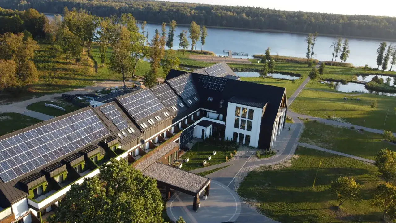 Nowoczesny hotel Mazury z panelami słonecznymi na dachu, położony nad jeziorem, otoczony zielenią.