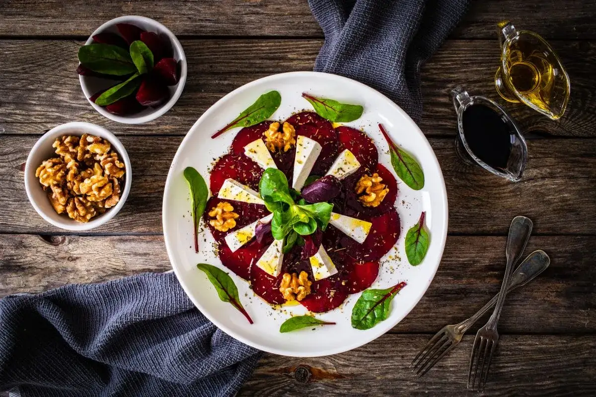 Eleganckie carpaccio z buraka z serem feta, orzechami włoskimi i świeżymi ziołami, podane na drewnianym stole.