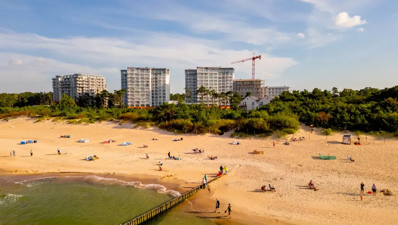 Piękna plaża przy Porta Mare Active & Spa Resort. Ludzie odpoczywają na piasku, w tle nowoczesne budynki i las.