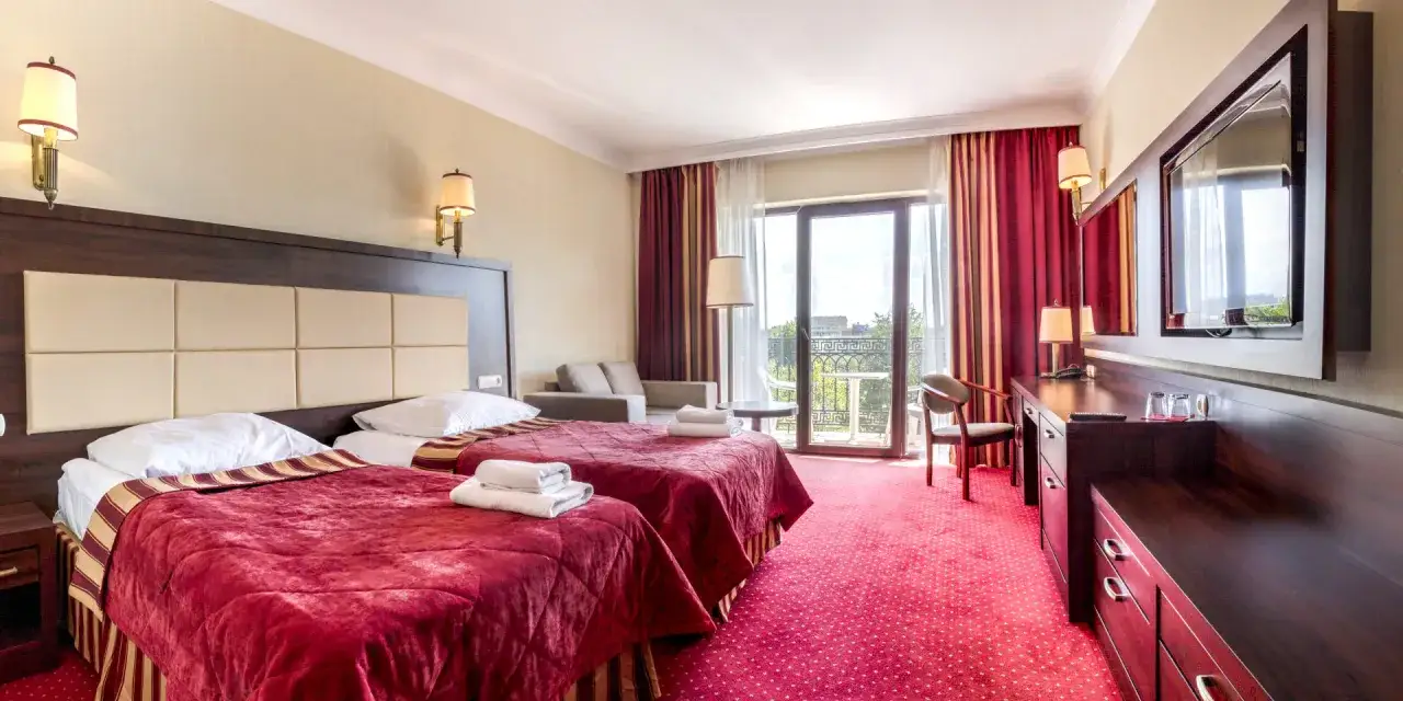 Pokój hotelowy w Doris Spa Kołobrzeg: dwa łóżka z czerwonymi narzutami, kanapa, balkon, TV.
