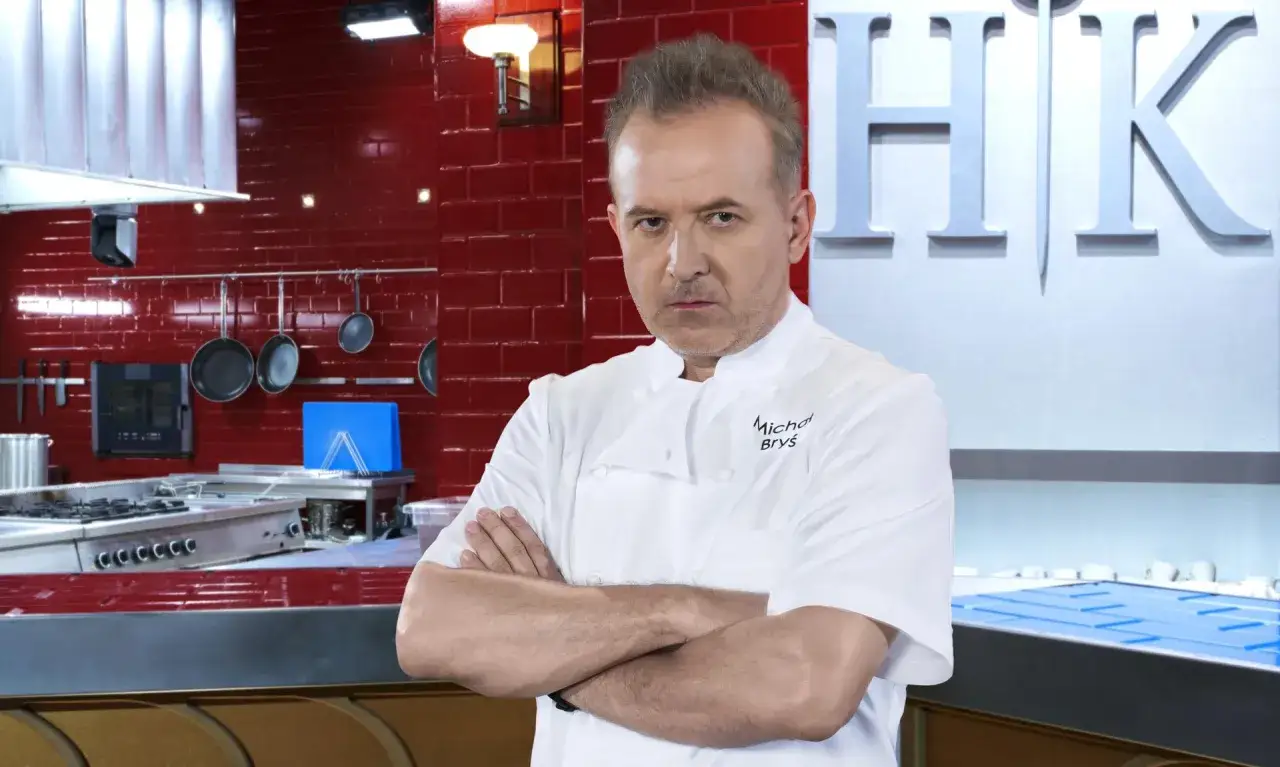 Michał Bryś w białym fartuchu, z rękami skrzyżowanymi na piersi, stoi przed profesjonalną kuchnią.